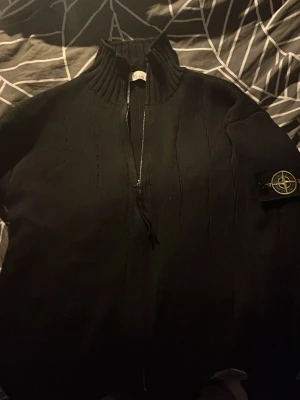 Svart ziptröja från Stone Island - Säljer en svart ziptröja från Stone Island med hög krage och ribbade detaljer. Tröjan har dragkedja framtill och den klassiska Stone Island-loggan på ärmen. Perfekt för dig som gillar streetwear och vill ha något stilrent och bekvämt.