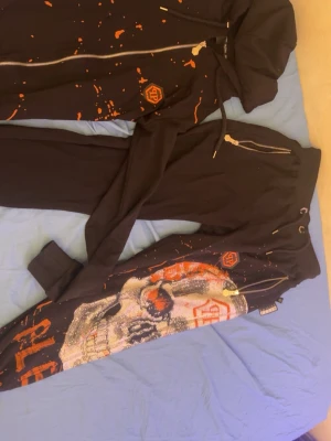 Philipp Plein med dödskalle dress svart/orange - Säljer ett par svarta mjukisbyxor från Philipp Plein med orangea färgstänk, dragkedjor och en stor dödskalle i strass på ena benet. Byxorna har dragsko i midjan och coola detaljer som sticker ut. Perfekta för dig som gillar streetwear och vill synas.