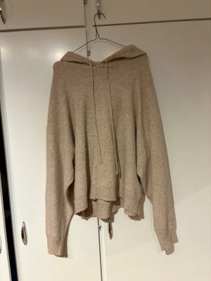 Beige stickad hoodie - Mysig beige stickad hoodie i oversized modell med huva och dragsko. Tillverkad i mjukt, stickat material som ger en avslappnad och chill vibe. Långa ärmar med ribbade muddar och något längre baktill för extra komfort.