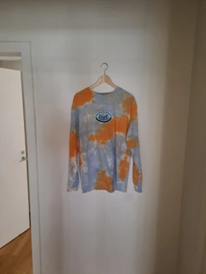 Tie-dye långärmad topp från HUF - Säljer en långärmad tie-dye topp från HUF med blå, orange och vita toner. Tröjan har ett stort HUF Worldwide-tryck på bröstet och är gjord i mjuk bomull. Perfekt för dig som gillar streetwear och vill sticka ut med färg.