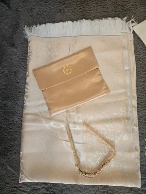 Mönstrad bönematta i satin, beige & blå - eleganta bönemattor i satin med vackert mönster och fransar i kanterna. En i ljusbeige och en i ljusblå, båda med matchande kuvertväska och dekorativt pärlband. Perfekt accessoar för att lyfta din outfit.