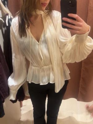 Krämvit blus med scarfdetalj - Snygg krämvit blus i glansigt material med v-ringning, volang nertill och tillhörande scarf vid halsen. säljer då den är förstor🩷 aldrig använd med lappen kvar💕