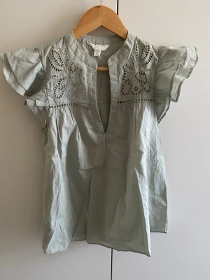 Ljusgrön blus med broderi från H&M - Söt ljusblå blus från H&M med broderade detaljer och hålmönster upptill. Blusen har volangiga korta ärmar och en liten ståkrage med v-ringning. Tillverkad i ett tunt, luftigt bomullstyg som passar perfekt till sommaren.