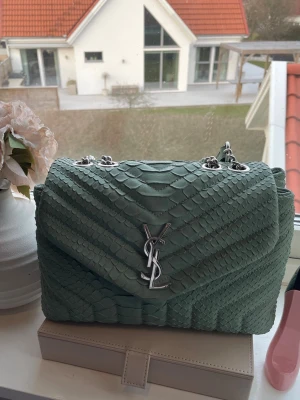 Grön YSL handväska i ormskinn - Snygg handväska från YSL i ljusgrönt ormskinnsmönstrat läder med silverfärgad kedjerem och ikonisk YSL-logga framtill. Väskan har en klassisk rektangulär form och stilren design som sticker ut.