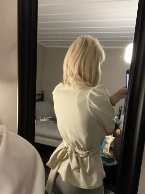 Krämvit omlottblus med knytband - Snygg krämvit omlottblus med korta ärmar och markerad midja tack vare ett knytband i ryggen. Blusen har en djup v-ringning och en lätt figurnära passform som ger en stilren och modern look. Perfekt att matcha med både jeans och kjol.