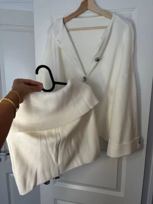 Vit kofta med stora knappar - Säljer en stilren vit kofta med stora silverfärgade knappar och breda ärmslut. Koftan har en lös passform och är tillverkad i ett mjukt stickat material. Perfekt för lager-på-lager och enkel att matcha med olika outfits.