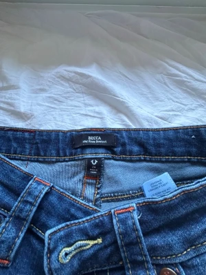 True Religion Becca Bootcut Jeans - Säljer ett par blå bootcut jeans från True Religion, modell Becca Mid Rise Bootcut. Jeansen har dekorativa fickor bak med stora knappar och kontrastsömmar. Klassisk femficksmodell i denim med snygga detaljer och logopatch bak.