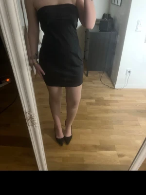 Svart axelbandslös kort klänning - Stilren svart axelbandslös klänning med knyt i ryggen. Kort modell som sitter tight och markerar figuren. Perfekt för dig som gillar minimalistisk och elegant stil.