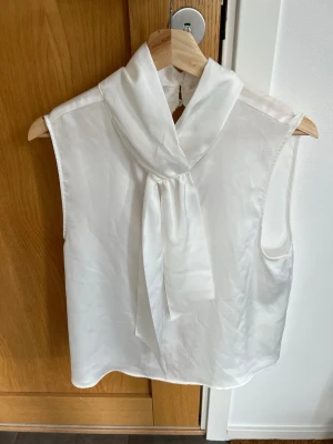 Vit ärmlös blus med knytkrage - Stilren vit blus i tunt, lätt material med hög krage och dekorativ knytning framtill. Blusen är ärmlös och har en öppning med knapp i nacken samt slits bak. Perfekt för en chic och modern look. Aldrig använd, helt ny💖! 