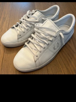Vita Fred Perry sneakers i läder - Säljer ett par stilrena vita sneakers från Fred Perry i läder med klassisk logga på sidan. Skorna har låga skaft, vita snören och diskreta sömmar för en clean look. Perfekta för dig som gillar minimalistisk och tidlös stil.