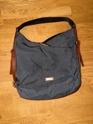 Mörkgrå axelväska med bruna detaljer - Snygg mörkgrå axelväska med bruna läderdetaljer och justerbar axelrem. Väskan har en mjuk, slouchy form och en liten metallbricka framtill. Perfekt för dig som vill ha en stilren och rymlig väska till vardagen.