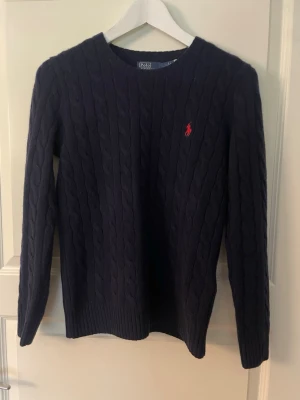 Mörkblå kabelstickad tröja Polo Ralph Lauren - Säljer en mörkblå kabelstickad tröja från Polo Ralph Lauren med klassisk rund halsringning och långärmad design. Tröjan har det ikoniska röda Polo-logot broderat på bröstet och är tillverkad i mjukt material som ger en skön känsla.