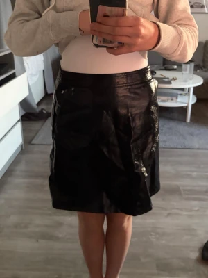 Svart glansig kjol i skinnimitation - Säljer en svart, glansig kortkjol i skinnimitation. Kjolen har en rak passform och sitter högt i midjan. Perfekt för dig som vill ha en edgy och trendig look. Inga synliga detaljer som dragkedja eller knappar på framsidan.