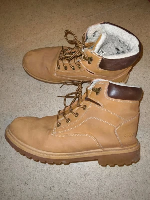  kängor med foder - Säljer ett par beige kängor med brunt läderdetalj vid ankeln och grov sula. Skorna har snörning med metallhål och är fodrade med mjukt, ljust material för extra värme. Perfekta för kalla dagar och har en robust design.