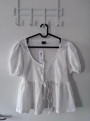 Vit puffärmad topp från Gina Tricot - Söt vit topp från Gina Tricot med puffärmar och volang nedtill. Toppen har en djup rundad urringning och knytband framtill. Tillverkad i ett lätt, strukturerat tyg som ger en luftig känsla. Perfekt för en trendig och avslappnad look.