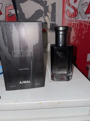 Ajmal Carbon Eau de Parfum 100ml - Ajmal Carbon Eau de Parfum är en parfym i en stilren svart glasflaska med metallfärgad kork. Doften kommer i en snygg svart kartong med silverdetaljer. Flaskan rymmer 100 ml och har en modern, maskulin design som passar perfekt på hyllan.