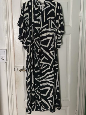 Svartvit mönstrad långklänning - Svart långklänning med vitt abstrakt mönster och små gulddetaljer. Klänningen har v-ringning, lös passform, volangärmar och knytband i midjan. Luftigt material som känns skönt mot huden och ger en avslappnad men trendig look.