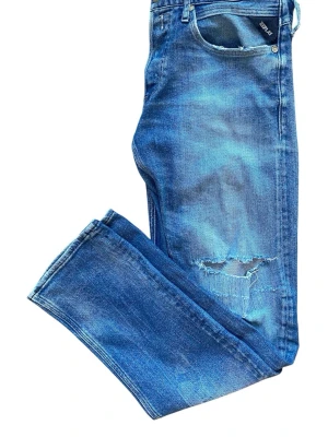 Schysta Replay jeans  - Säljer ett par grischiga blå jeans från Replay  med slitningar och hål på ena benet. Jeansen har klassisk femficksmodell, raka ben och ljusa tvättade partier för en trendig look. Perfekta för dig som gillar en avslappnad och cool stil.