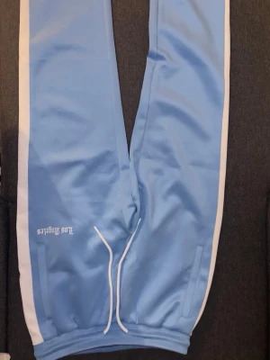 Blå mjukisbyxor från Black Squad - Snygga ljusblå mjukisbyxor från Black Squad med vita sidoränder och dragsko i midjan. Tillverkade i ett glansigt, sportigt material som känns riktigt skönt. Diskret tryck på ena benet och muddar vid bensluten för en clean look.