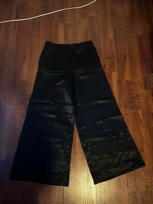 Svarta vida byxor i satin - Säljer ett par svarta byxor från ARKET med breda ben i glansigt silkesmaterial!  Byxorna är mid-waist och en enkel design utan synliga detaljer. .
