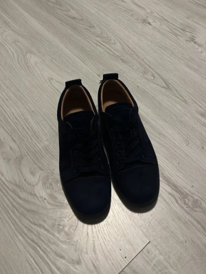 Mörkblå sneakers från Christian Louboutin - Snygga mörkblå sneakers från Christian Louboutin med klassisk röd sula och diskret logga undertill. Skorna har en låg profil, rund tå och är tillverkade i mocka med matchande mörkblå snörning. Perfekta för dig som gillar exklusiv och stilren design.
