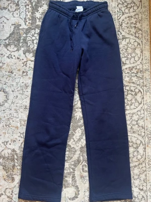 Mörkblå mjukisbyxor från Gina - Mörkblå mjukisbyxor med rak passform och resår i midjan samt dragsko för justerbar komfort. Tillverkade i mjukt och skönt material, perfekta för chill dagar. Klassisk enkel design utan synliga loggor eller detaljer.