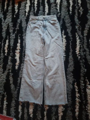 Ljusblåa vida jeans med rå kant - Säljer ett par ljusblå jeans med hög midja och raka, vida ben. Jeansen har råa, avklippta benslut och klassiska fem fickor. Perfekta för en avslappnad och trendig look. Materialet är denim i bomull.