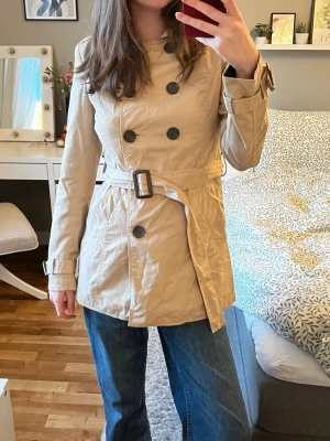 Beige trenchcoat med bälte - Stilren beige trenchcoat med dubbelknäppning och bälte i midjan. Klassisk krage och knappar framtill. Jackan har långa ärmar och justerbara spännen vid ärmsluten. Perfekt för en trendig och tidlös look.