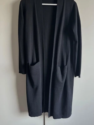 Svart lång Cardigan med fickor  - Stilren svart lång cardigan med öppet fall och två stora fickor framtill. Koftan har långa ärmar och är gjord i ett mjukt, stickat material som känns skönt mot huden. Perfekt att slänga över en t-shirt eller topp för en avslappnad look.