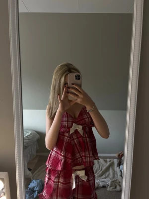 Röd- och vitrutigt pyjamasset - Supersött pyjamasset med röd- och vitrutigt mönster och två stora cremefärgade rosetter framtill. Setet har volanger och tunna axelband, perfekt för mysiga kvällar hemma. Materialet känns lätt och luftigt, perfekt för sommarnätter.