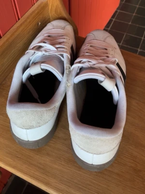 Vita Adidas sneakers med svarta ränder - Snygga vita Adidas sneakers med klassiska svarta ränder på sidorna och gummisula. Ovandelen är i läder med detaljer i mocka på hälen och tån. Skorna har lågt skaft och vita skosnören. Perfekta för en sportig och stilren look.