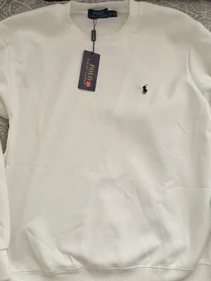 Vit sweatshirt från Polo Ralph Lauren - Klassisk vit sweatshirt från Polo Ralph Lauren med rund halsringning och den ikoniska broderade loggan på bröstet. Tröjan har ribbade muddar vid ärmslut och nederkant. Perfekt för en clean och stilren look.