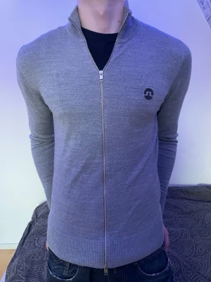 Grå ziptröja med hög krage - Snygg grå ziptröja från J.Lindeberg med hög krage och hel dragkedja framtill. Tröjan har ribbade muddar och nederkant samt en diskret logga på bröstet. Perfekt till jeans eller chinos för en clean look.