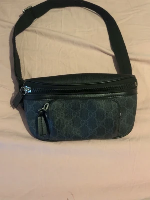 Svart magväska från ##### - Snygg svart magväska från ##### med klassiskt GG-mönster i tyg och detaljer i läder. Väskan har justerbart band, två dragkedjefack och silverfärgade metalldetaljer. Perfekt för att bära det viktigaste på ett stilrent sätt.