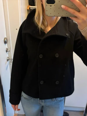 Svart kort dubbelknäppt rock - Stilren svart rock med dubbelknäppning och bred krage. Modellen är kortare och har en klassisk, rak passform. Perfekt för dig som gillar en tidlös look. Rocken har stora svarta knappar och är troligtvis i ullblandning. Använd 3 gånger, nypris 999 kr