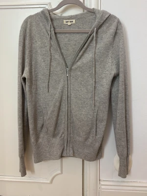 Grå hoodie i kashmir från Soft Goat - Super populär,mjuk och stilren grå hoodie från Soft Goat i kashmir. Dragkedja framtill, två fickor och justerbar huva med snören. Perfekt för en chill och avslappnad look. Ribbstickade muddar vid ärmslut och nederkant. Köpt för 2295kr online 