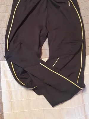 Svarta träningsbyxor med gula detaljer - Säljer ett par svarta träningsbyxor från Craft med tunna gula linjer längs benen och små reflekterande prickar vid höften. Byxorna är i ett stretchigt funktionsmaterial som passar perfekt till löpning eller gym. Snygg och sportig design! Storlek L/12-14.