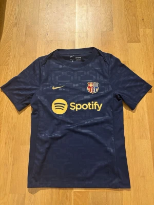 Blå FC Barcelona tröja Nike - Snygg mörkblå FC Barcelona matchtröja från Nike med klubbmärke och Spotify-logga på bröstet. Tröjan har korta ärmar, rund hals och är tillverkad i ett lätt, ventilerande funktionsmaterial. Perfekt för fotbollsträning eller att bära till vardags. Ser ut och känns som ny. Den är endast använd en gång, vilket är varför prislappen ej är kvar. Skicka gärna vid frågor. 