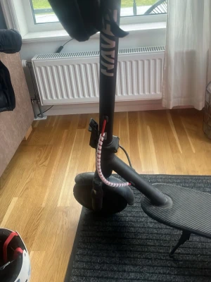 Svart elscooter med digital display - Säljer en svart elscooter från naver med digital display och röda detaljer på kablarna. Scootern har en bred fotplatta med räfflat mönster för bra grepp och en spiralformad låskabel runt styret. Perfekt för smidig transport i stan.