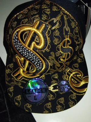 Svart keps med gulddetaljer från Leader - Säljer en svart keps från Leader med stora broderade dollartecken i guld och blingade stenar på framsidan. Kepsen har platt skärm och är täckt av små guldfärgade dollartryck. Perfekt för dig som vill sticka ut och gillar streetwear.