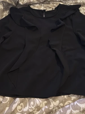 Svart blus med volangdetaljer - Stilren svart blus med långa ärmar och volangdetaljer framtill. Blusen har rund halsringning och resår vid ärmsluten för en snygg passform. Perfekt för dig som gillar en trendig och elegant look. Storlek 164. 