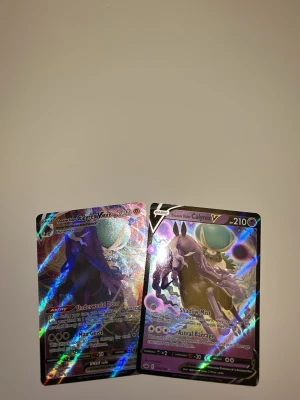 Pokémonkort Shadow Rider Calyrex VMAX - Två Pokémonkort med motiv av Shadow Rider Calyrex V och VMAX. Korten har holografisk yta med färger i blått, lila och svart samt detaljer i silver. Perfekt för samlare eller Pokémonfans som vill utöka sin kortsamling.