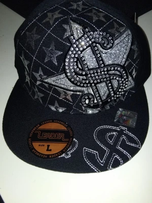 Svart keps med bling och dollartecken - Svart keps från Leader Headwear med stora glittriga dollar- och stjärnmotiv framtill. Kepsen har broderade detaljer, blingstenar och ett rutmönster i silver. Perfekt för dig som vill sticka ut med en unik och flashy stil.