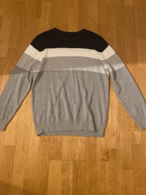 Grå stickad tröja - Snygg stickad tröja i grått med breda blockränder i svart och vitt upptill. Tröjan har rund halsringning och ribbade muddar vid ärmslut och nederkant. Jätte skönt material och är lent mot huden. Använd en gång!!!