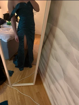 Mörkgrön axelbandslös balklänning - Snygg mörkgrön axelbandslös balklänning med volang upptill och hög slits framtill. Klänningen har figurnära passform och är gjord i ett glansigt tyg som ger en lyxig känsla. Perfekt för dig som vill sticka ut på nästa stora tillställning.