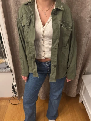 Grön overshirt med nitar - Stilren grön overshirt med coola nitar på axlar och bröstfickor. Jackan har en avslappnad passform, klassisk krage och stora fickor framtill. Perfekt att slänga över en topp för en casual look.