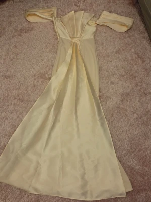 Champagnefärgad lång klänning i satin - Elegant långklänning i champagnefärgad satin med offshoulder-ärmar och veckad detalj framtill. Klänningen har en vacker, svepande kjol och en lyxig känsla som passar perfekt till speciella tillfällen.