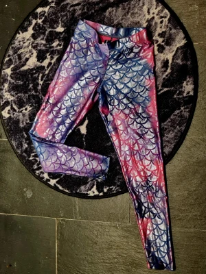 Black Milk mermaid leggings *limited edition* - Säljer ett par glansiga leggings, av märket Black Milk, i metallic med mermaidmönster i blått, rosa och lila. Limiterad utgåva!! Passa på!! Knappt använda pga fel storlek. Normal storlek M. De har hög midja och stretchigt material som sitter tajt. Perfekta för dig som vill sticka ut med en unik och färgstark stil.
