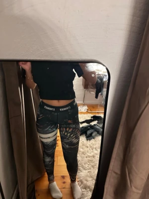 Mönstrade leggings från Stronger - Säljer ett par snygga leggings från Stronger med hög midja och elastisk linning där loggan syns tydligt. De har ett coolt abstrakt mönster i svart, vitt, beige och orange toner. Materialet är stretchigt och passar perfekt för träning eller chill.