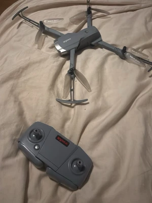Syma 30x grå drönare med kontroll - Säljer en grå drönare från Syma med tillhörande fjärrkontroll. Drönaren har fyra propellrar, vikbara armar och en inbyggd kamera undertill och en fram. Tillverkad i plast och designad för enkel styrning och smidig transport. Perfekt för dig som vill testa på att flyga drönare. Den har endast kört försiktigt.